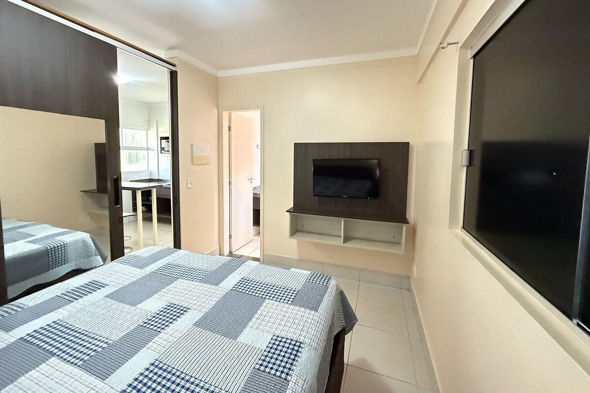 Un dormitorio con cama, televisor montado en la pared, espejo y puerta que da al baño.