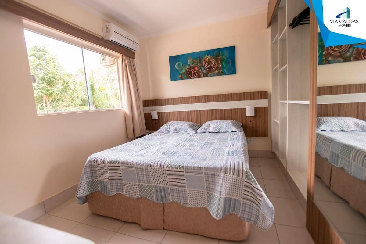 A bedroom with a bed, a window, an air conditioner, and a painting on the wall.