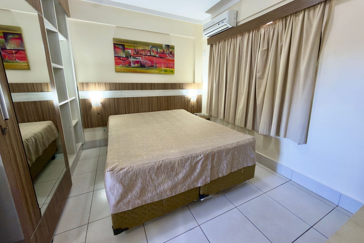 A hotel room with a bed, a painting on the wall, a window with curtains, and an air conditioning unit.