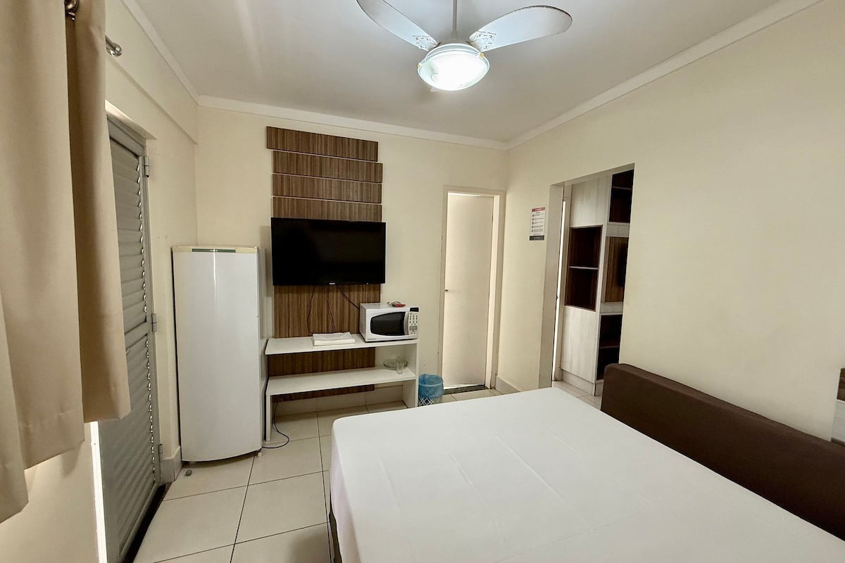 A hotel room with a bed, a television mounted on the wall, a microwave, a refrigerator, a ceiling fan, and a door leading to another room.