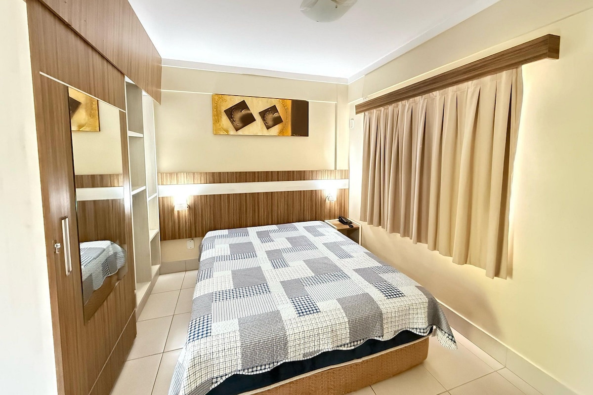 A bedroom with a bed, a wardrobe, a mirror, a bedside table, a wall-mounted light, and a window with curtains.