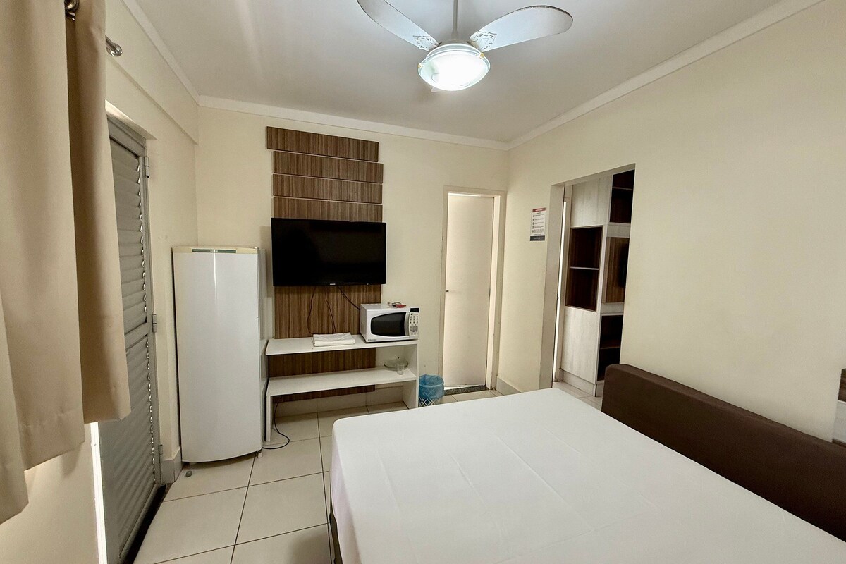 Habitación de hotel con una cama, televisión montada en la pared, microondas, refrigerador, ventilador de techo y una puerta que da a otra habitación.