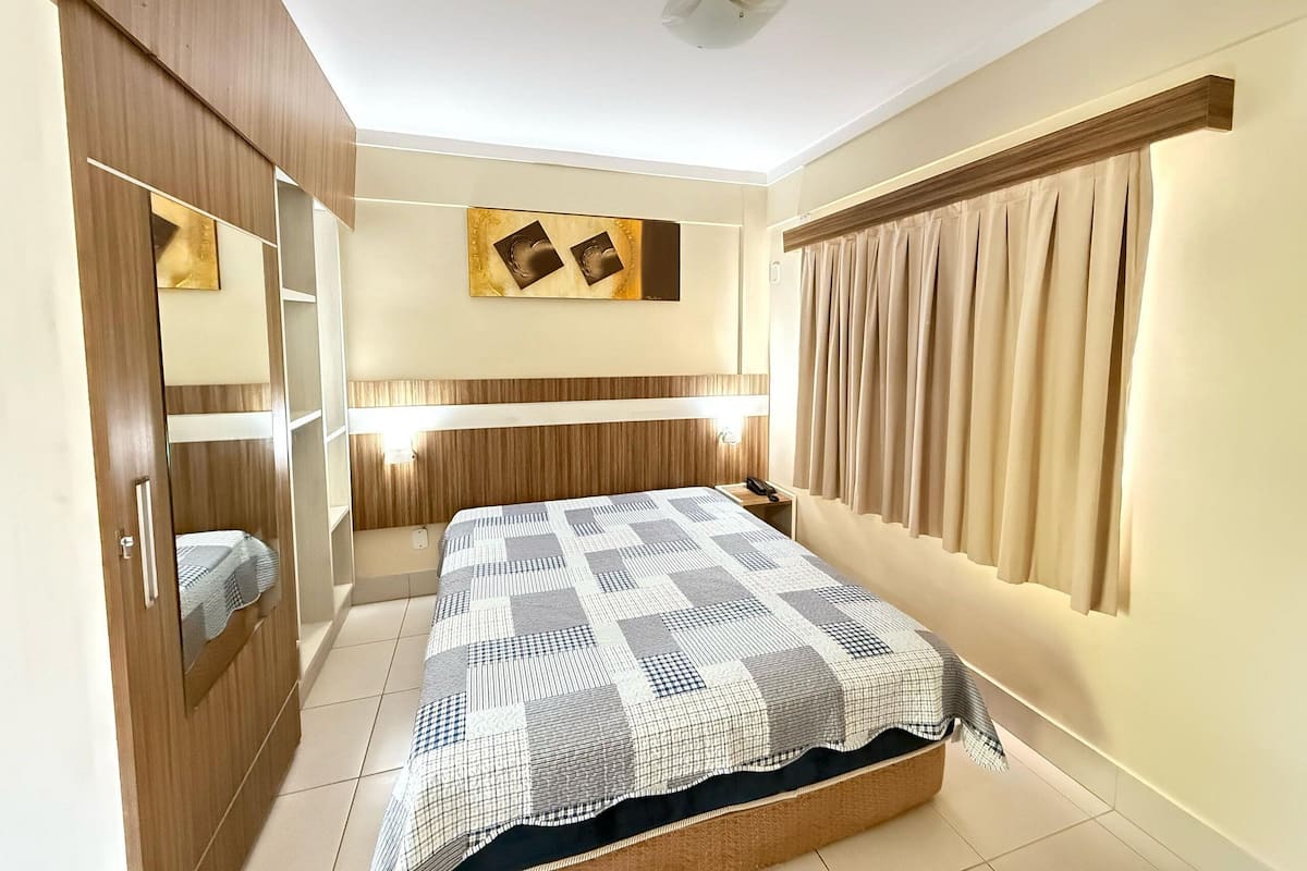 A bedroom with a bed, a wardrobe, a mirror, a bedside table, a wall-mounted light, and a window with curtains.