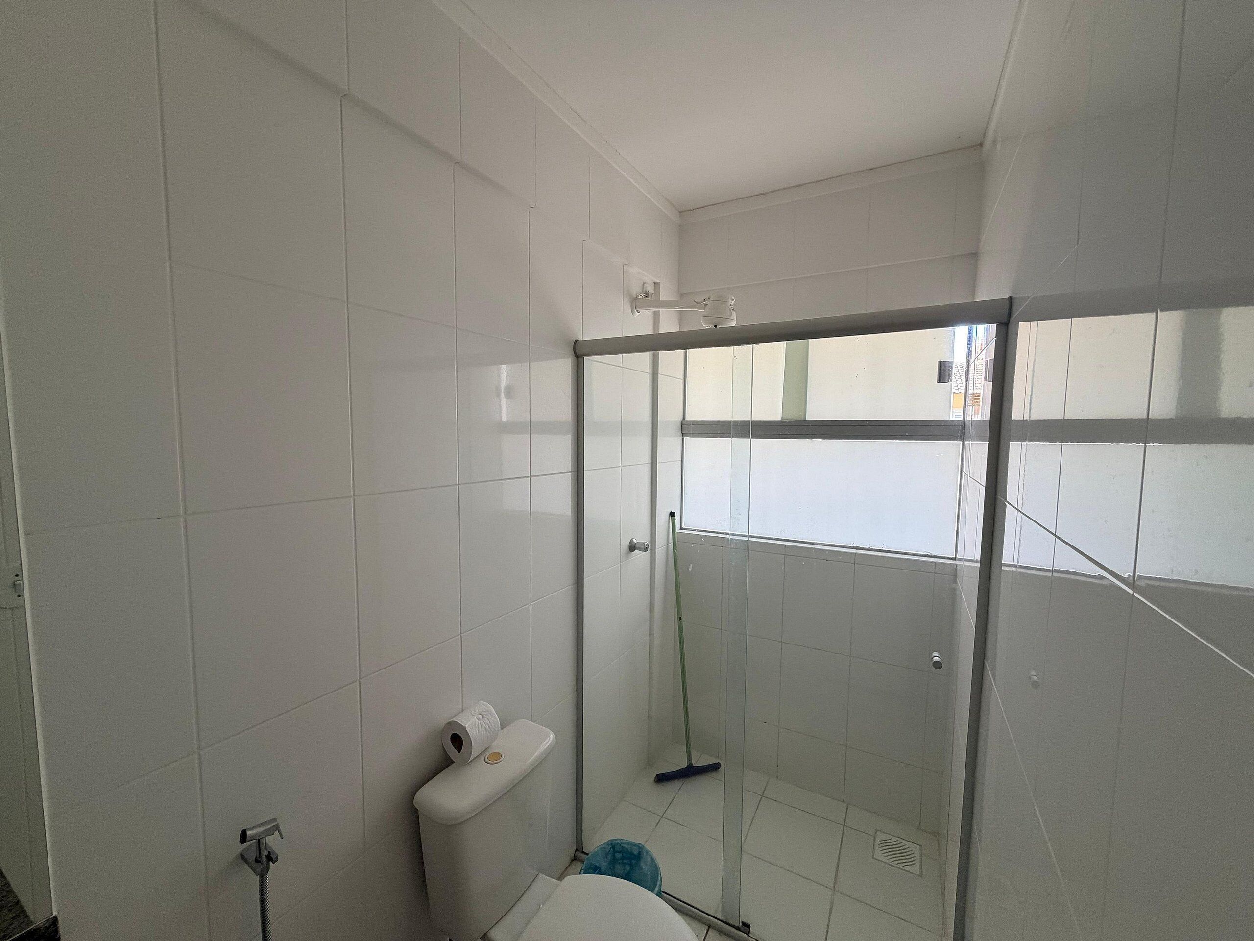A bathroom with white tiled walls, a glass shower enclosure, a toilet, and a mop leaning against the wall.