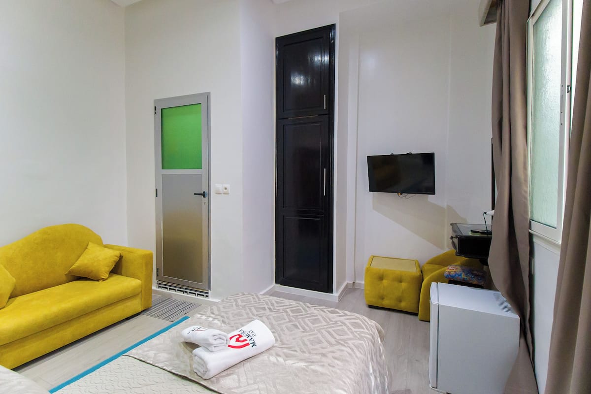 Una habitación de hotel con un sofá amarillo, una cama con colcha estampada, una mesita de noche blanca, un pequeño otoman amarillo y un televisor montado en la pared.