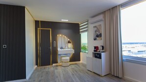 A modern hotel room with a wooden floor, a vanity with a mirror, a small stool, a minibar, and a view of a marina through large windows. - MAMORA BAY (Tangier)