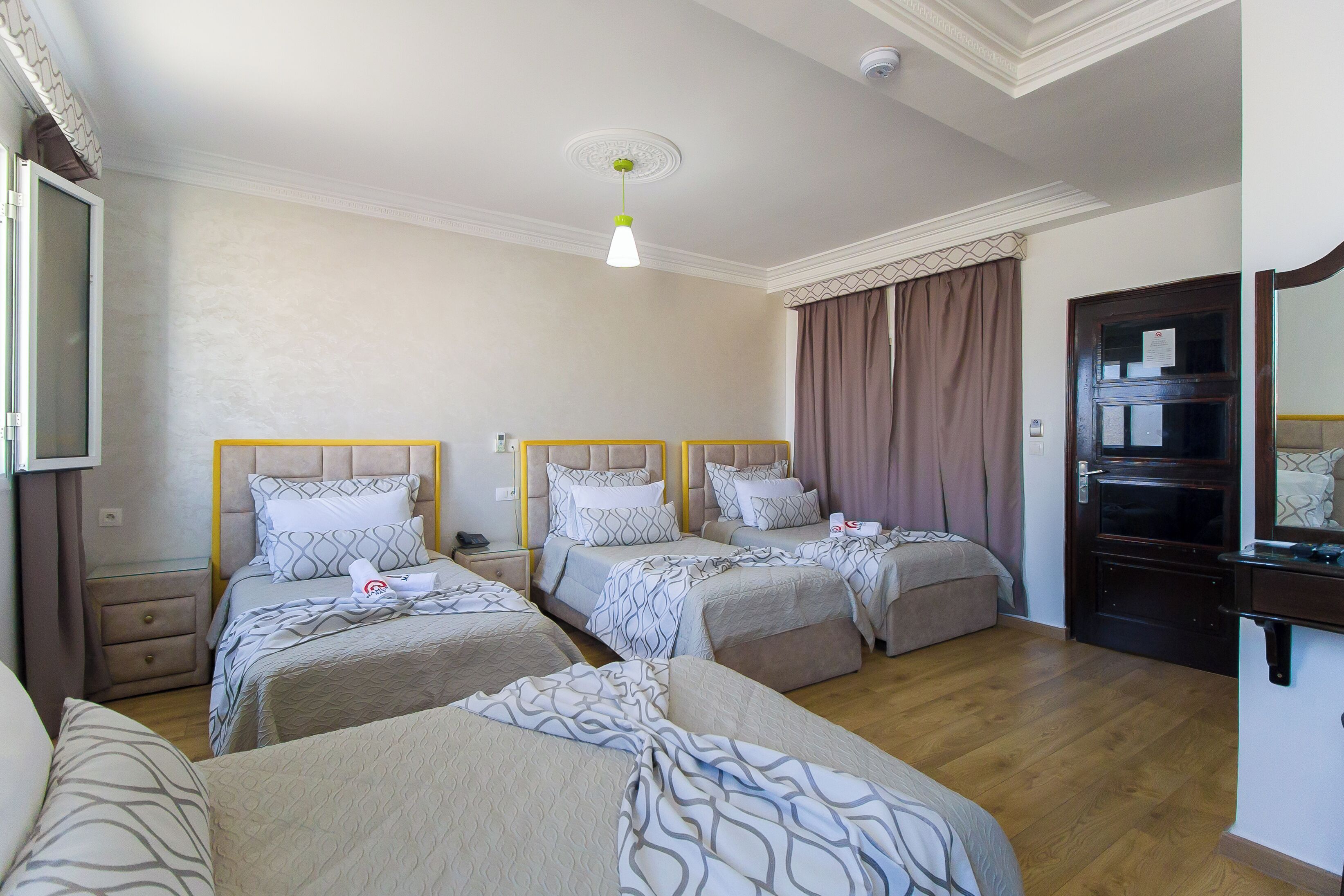 Quarto de hotel com três camas, piso de madeira, luminária no teto e porta de madeira escura.