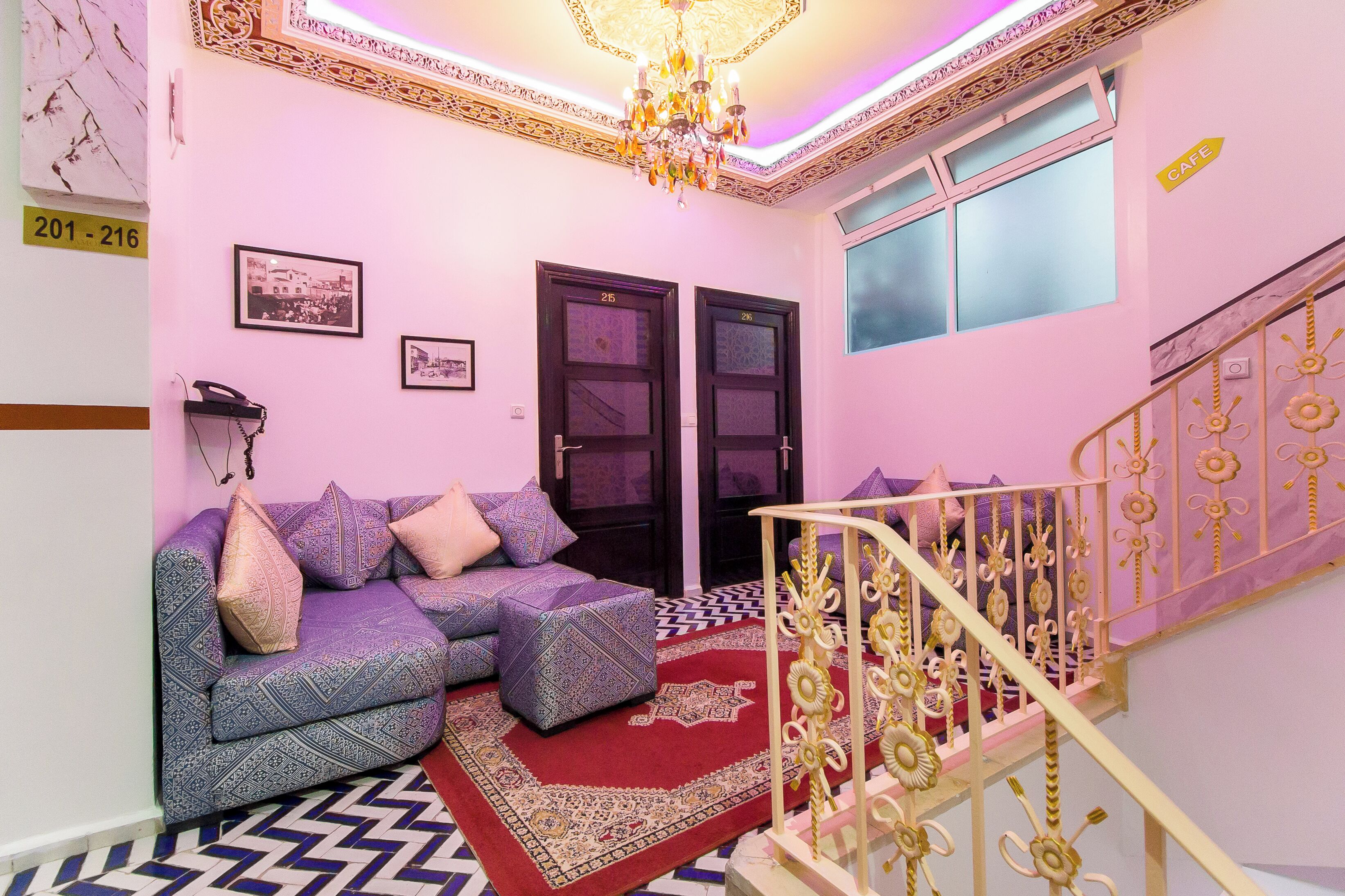 Une pièce avec un canapé violet, un tapis aux motifs rouges et dorés, un lustre et un escalier doté de rampes décoratives.