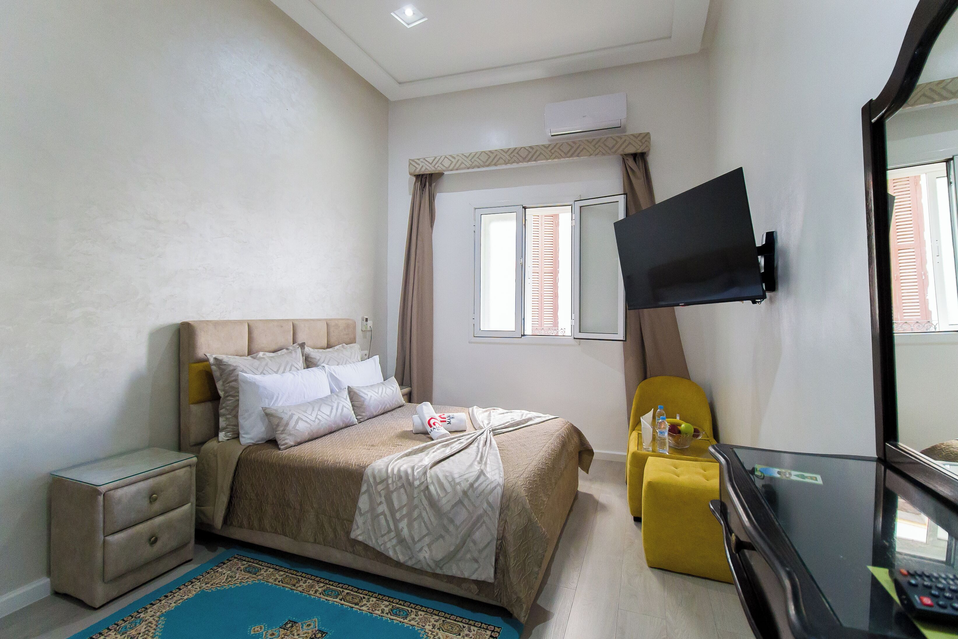 A hotel room with a bed, a nightstand, a TV mounted on the wall, a small yellow ottoman, and a window with curtains.