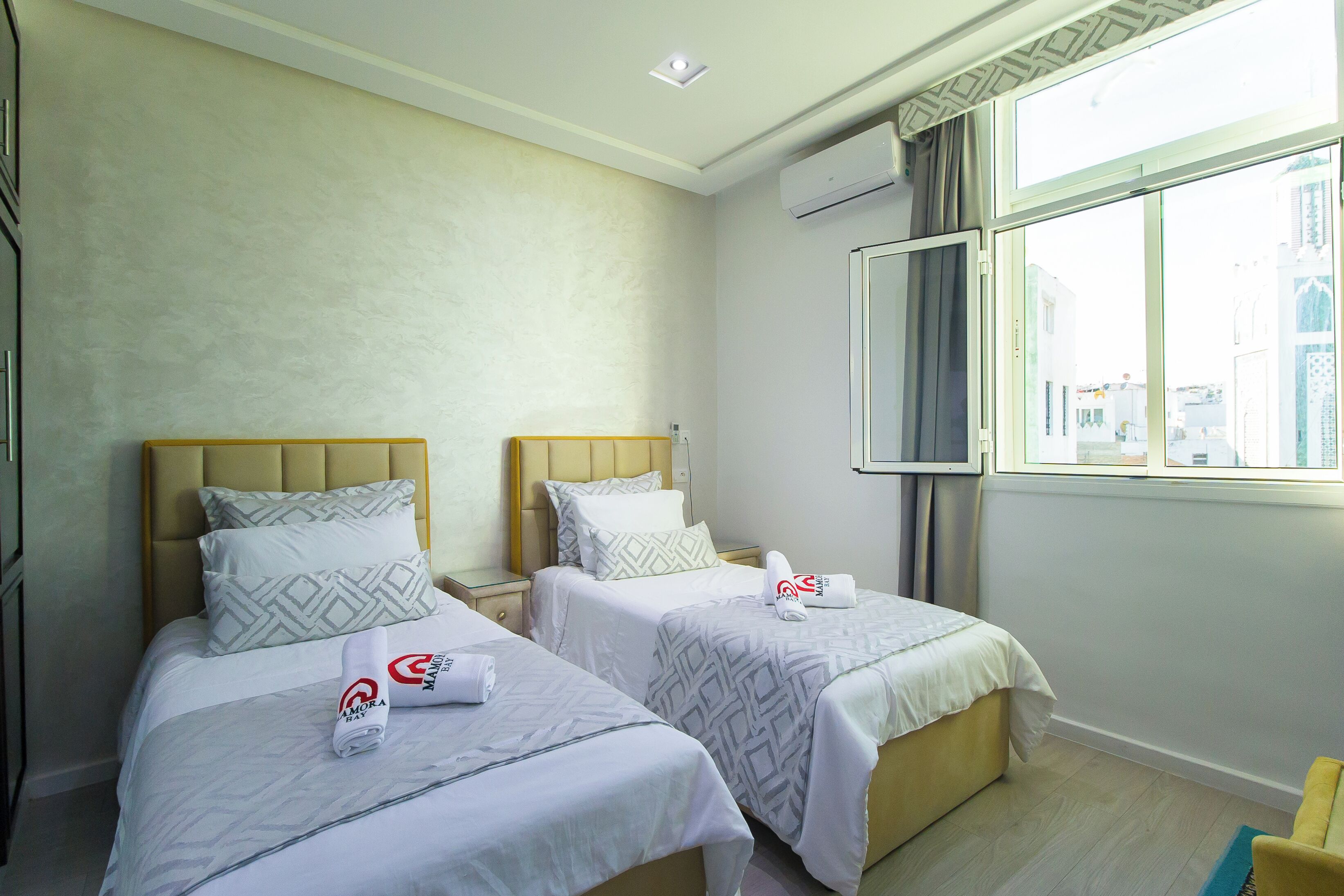 Una habitación de hotel con dos camas, cada una con cabecera y almohadas, una ventana con cortinas y un equipo de aire acondicionado en la pared.