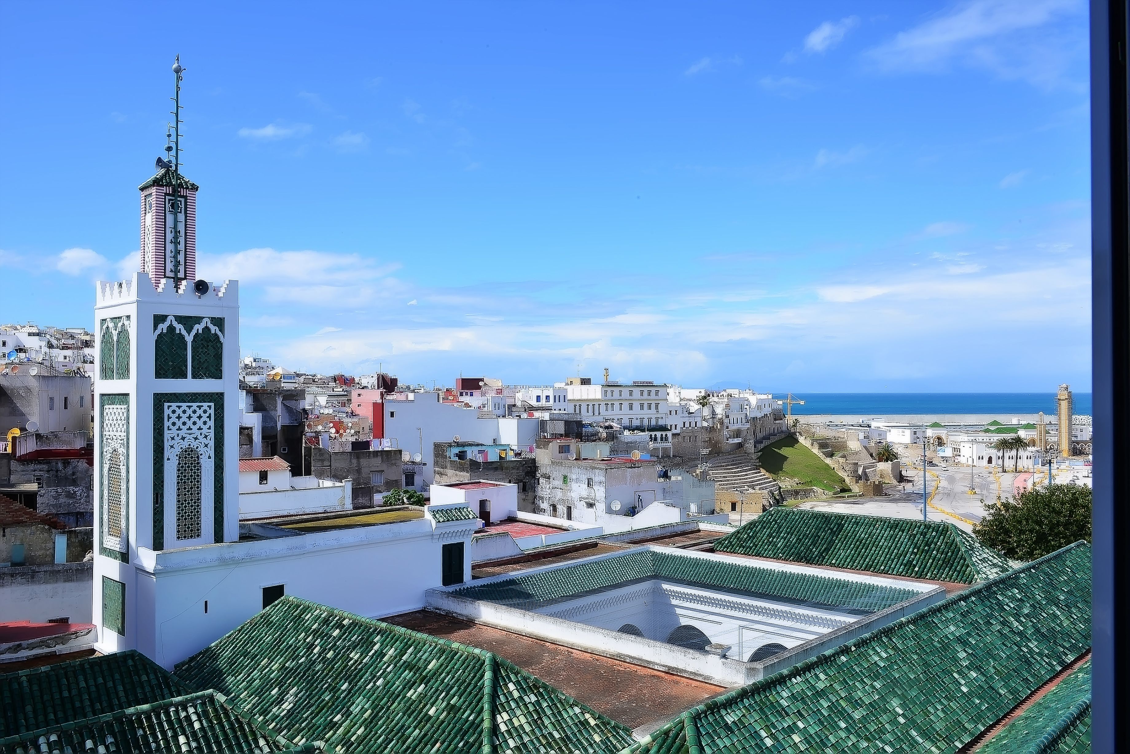 A view of a coastal town with a prominent white tower with green accents, a mosque with a green roof, and a clear blue sky.
