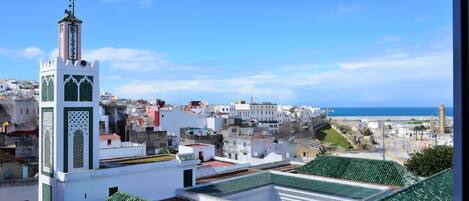 A view of a coastal town with a prominent white tower with green accents, a mosque with a green roof, and a clear blue sky.
