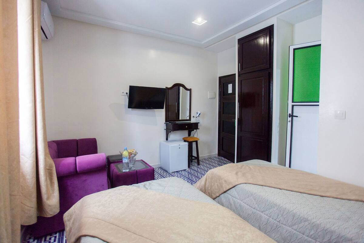 Habitación de hotel con dos camas, un sillón púrpura, una mesita con una botella y vasos, un televisor, un espejo y un armario.