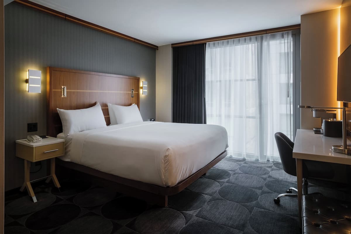 Ein Hotelzimmer mit einem großen Bett, einem Schreibtisch und einem Stuhl. Auf dem Nachttisch befindet sich ein Telefon und auf dem Schreibtisch eine Lampe.