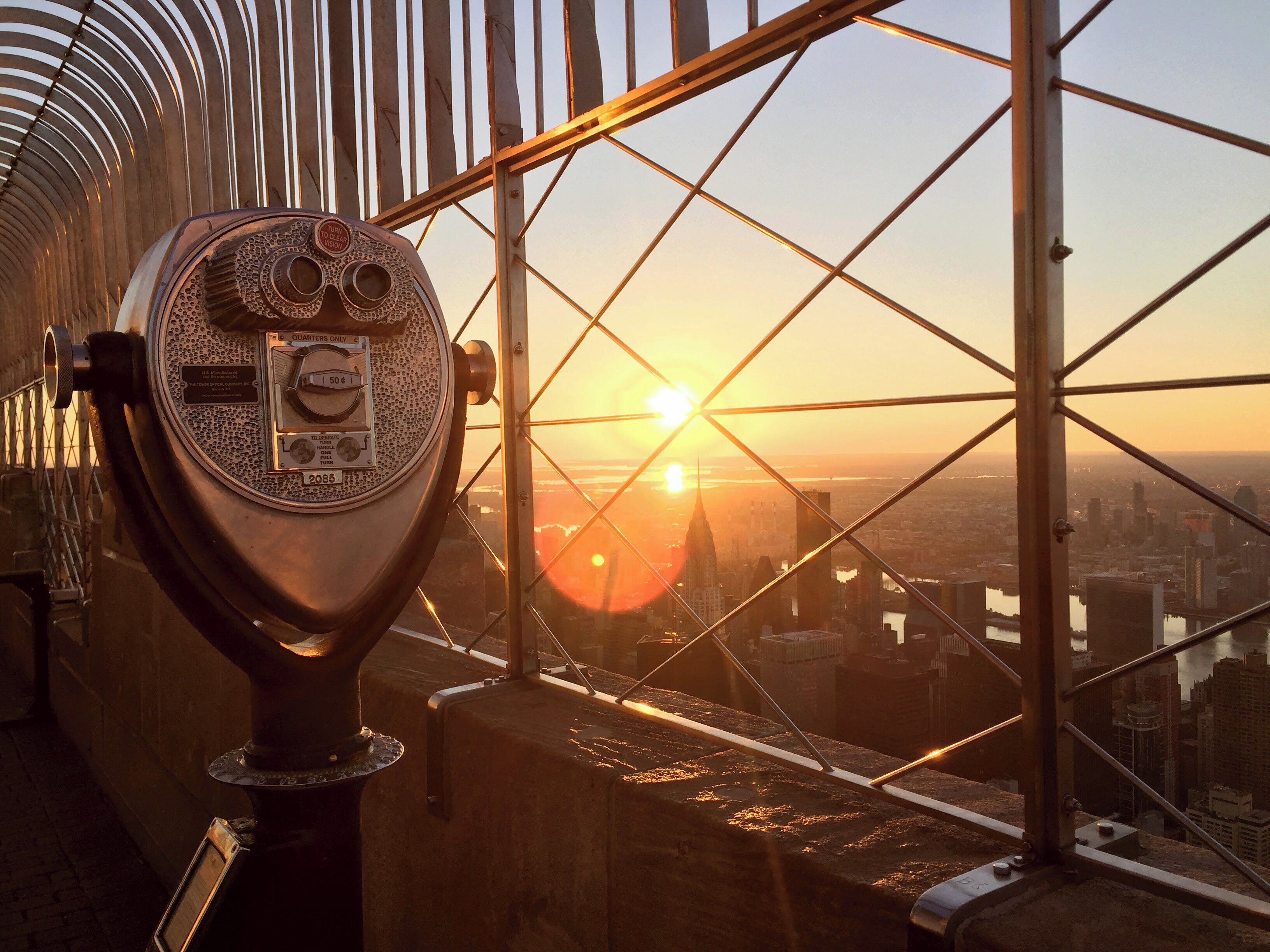 Un télescope ancien, agrémenté d’une plaque commémorative, est placé près d’une grande fenêtre donnant sur un paysage urbain au coucher du soleil.