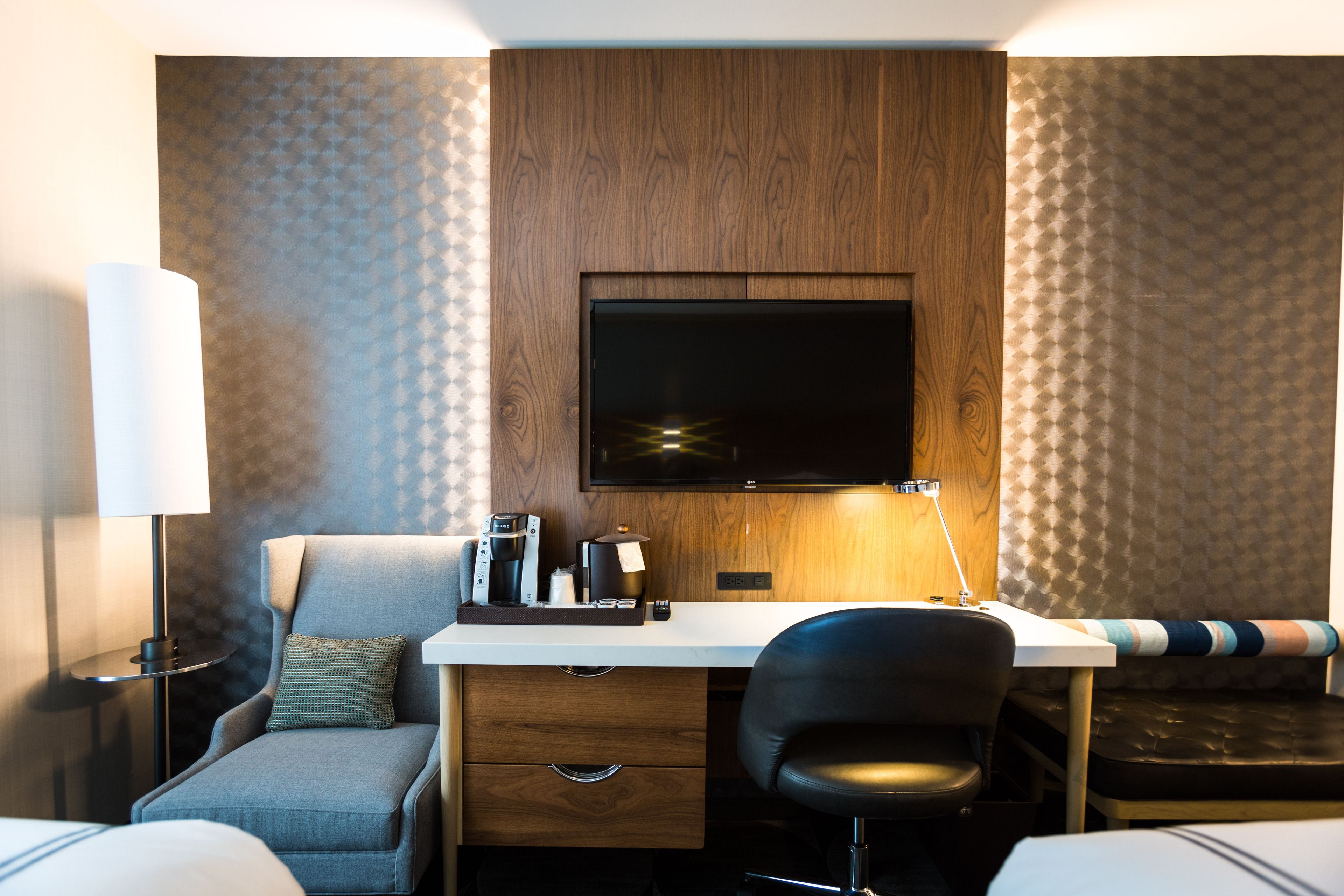 A hotel room with a wooden panel wall, a flat-screen TV, a desk with a lamp, a chair, a bed, and a sofa.