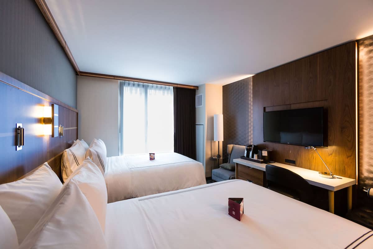 Ein Hotelzimmer mit zwei Betten, einem Schreibtisch, einem Fernseher und einem Fenster mit Vorhängen.