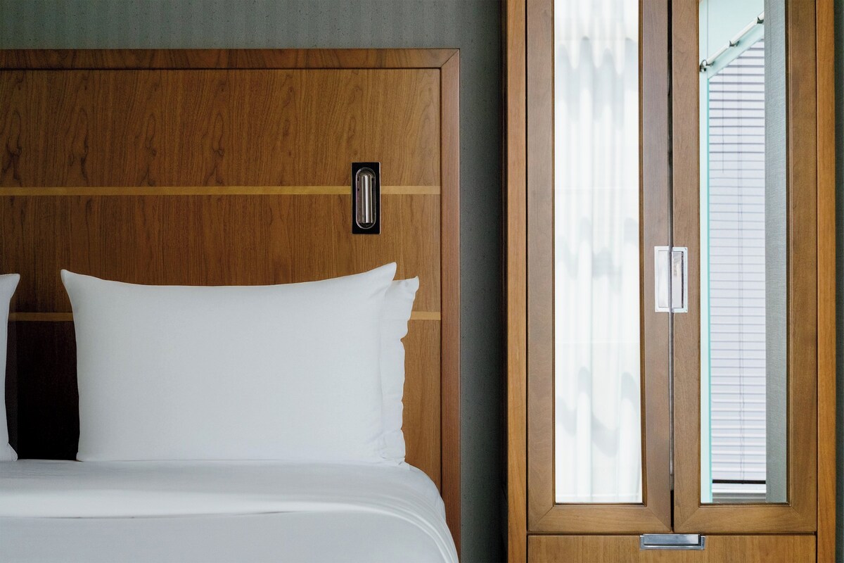 Cabecera de madera con luz integrada, colcha blanca y vistas a una ventana con persianas.