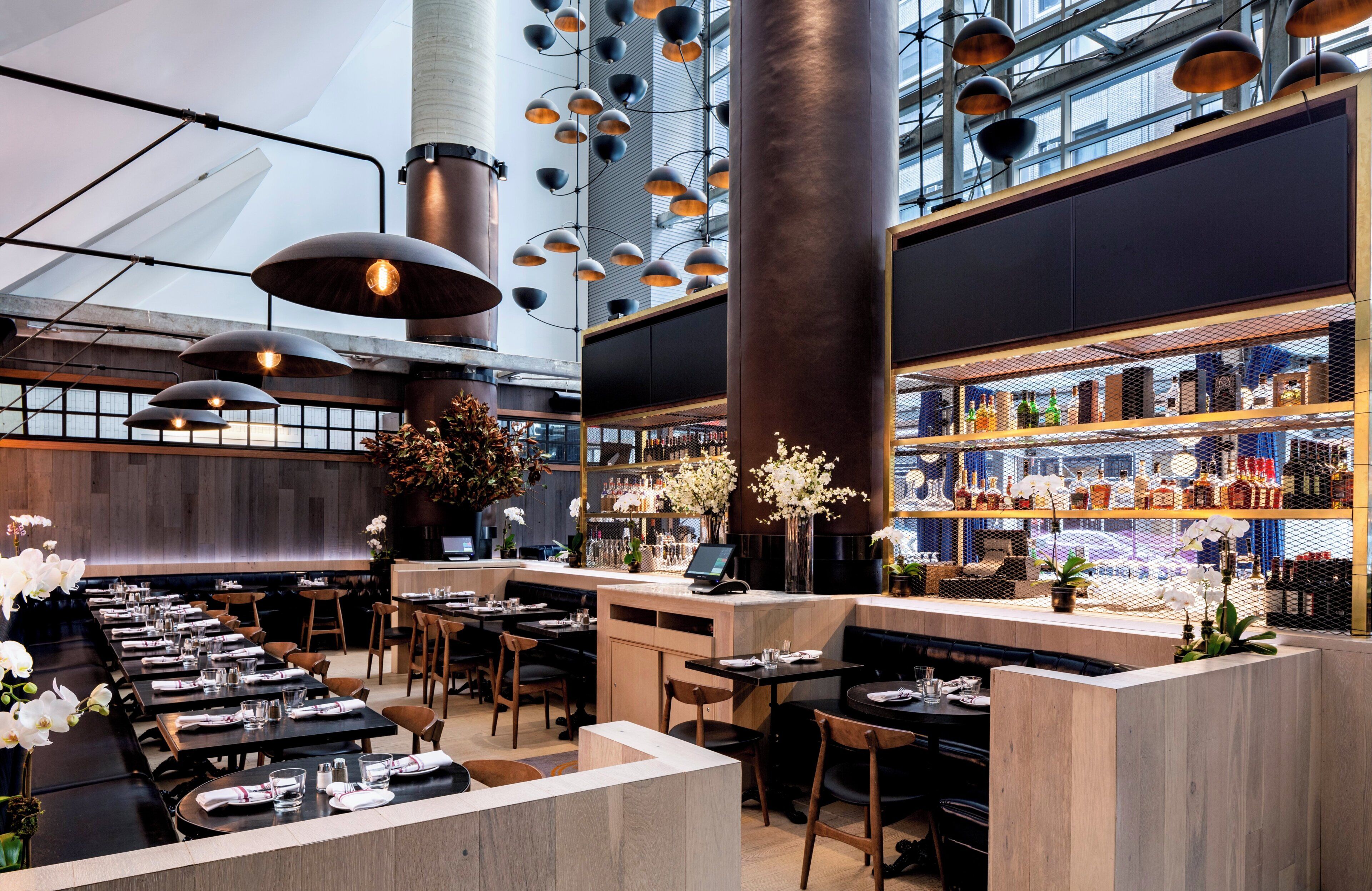 Un restaurant moderne avec un intérieur caractérisé par de hauts plafonds, des conduits apparents et un bar doté d’un présentoir à bouteilles de vin.