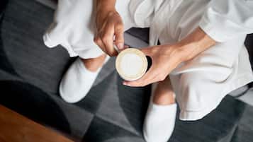 Una persona sosteniendo una taza de café, vestida con ropa blanca, sentada sobre un suelo con diseño.