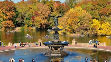 A public park with a central fountain, surrounded by a lake and autumn-colored trees.