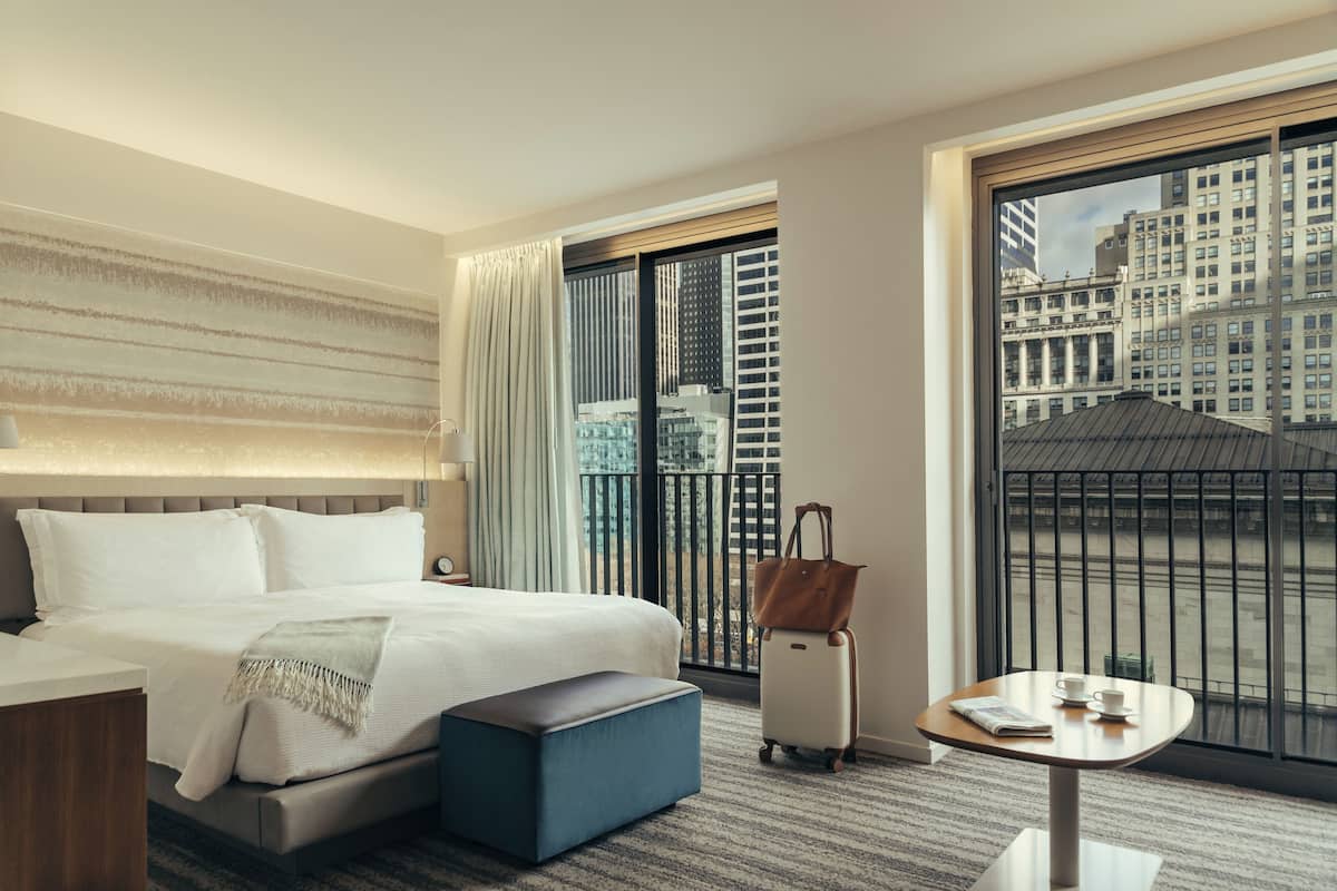 Habitación de hotel con una cama grande, una mesita con una taza y un periódico, una maleta y vistas a un paisaje urbano a través de un ventanal amplio.