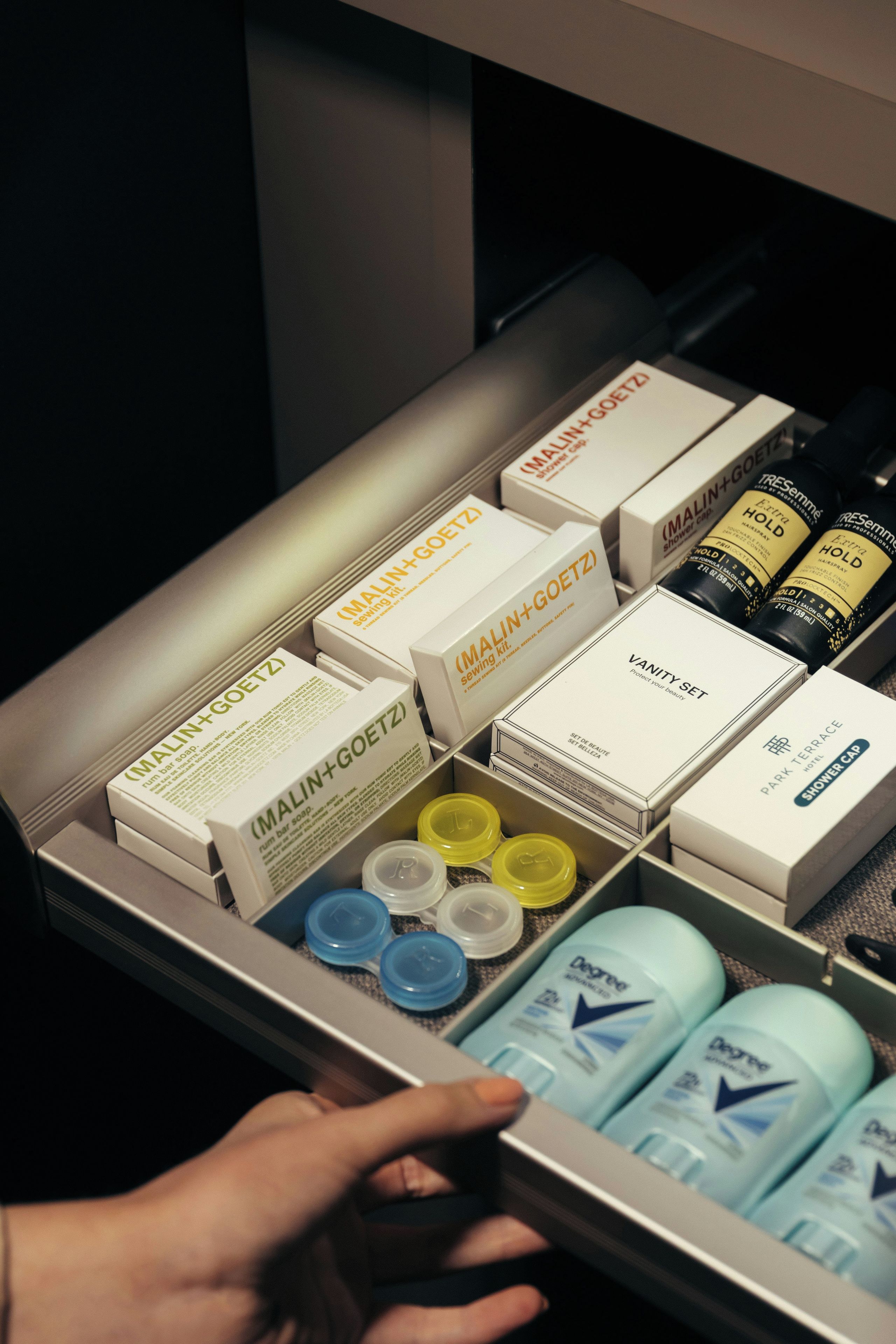 A drawer containing various toiletries including a Malin+Goetz bar soap, a shower cap, and a set of beauty products.