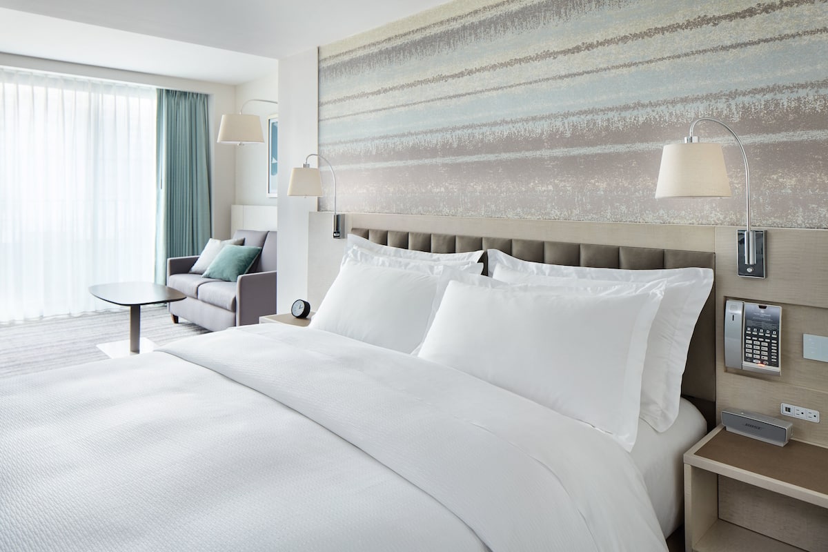 Habitación de hotel con una cama grande, una mesita de noche, una lámpara, una silla y vistas al exterior.