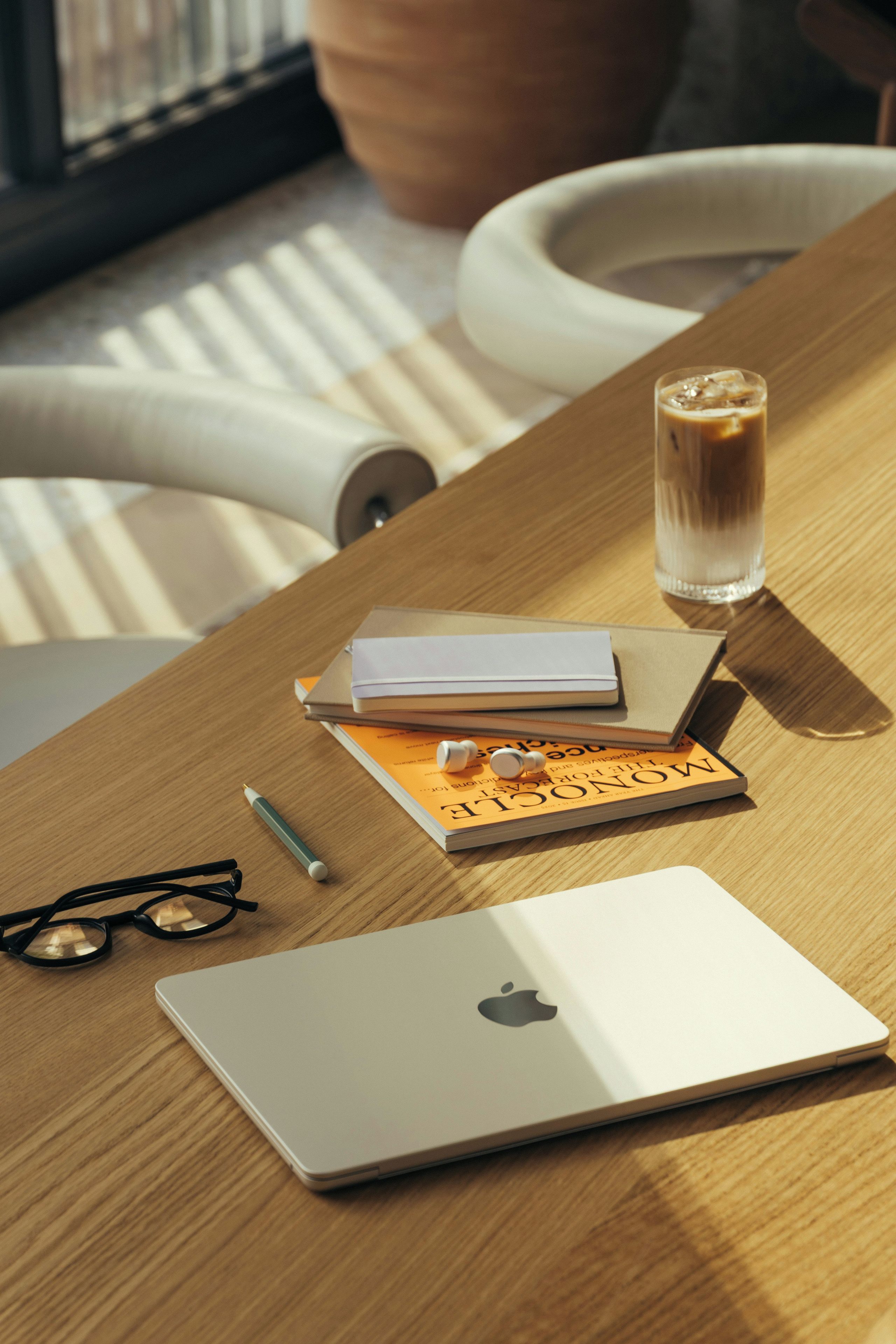 A wooden table with a laptop, glasses, a pen, a notebook, a glass of water, and a pair of earphones.