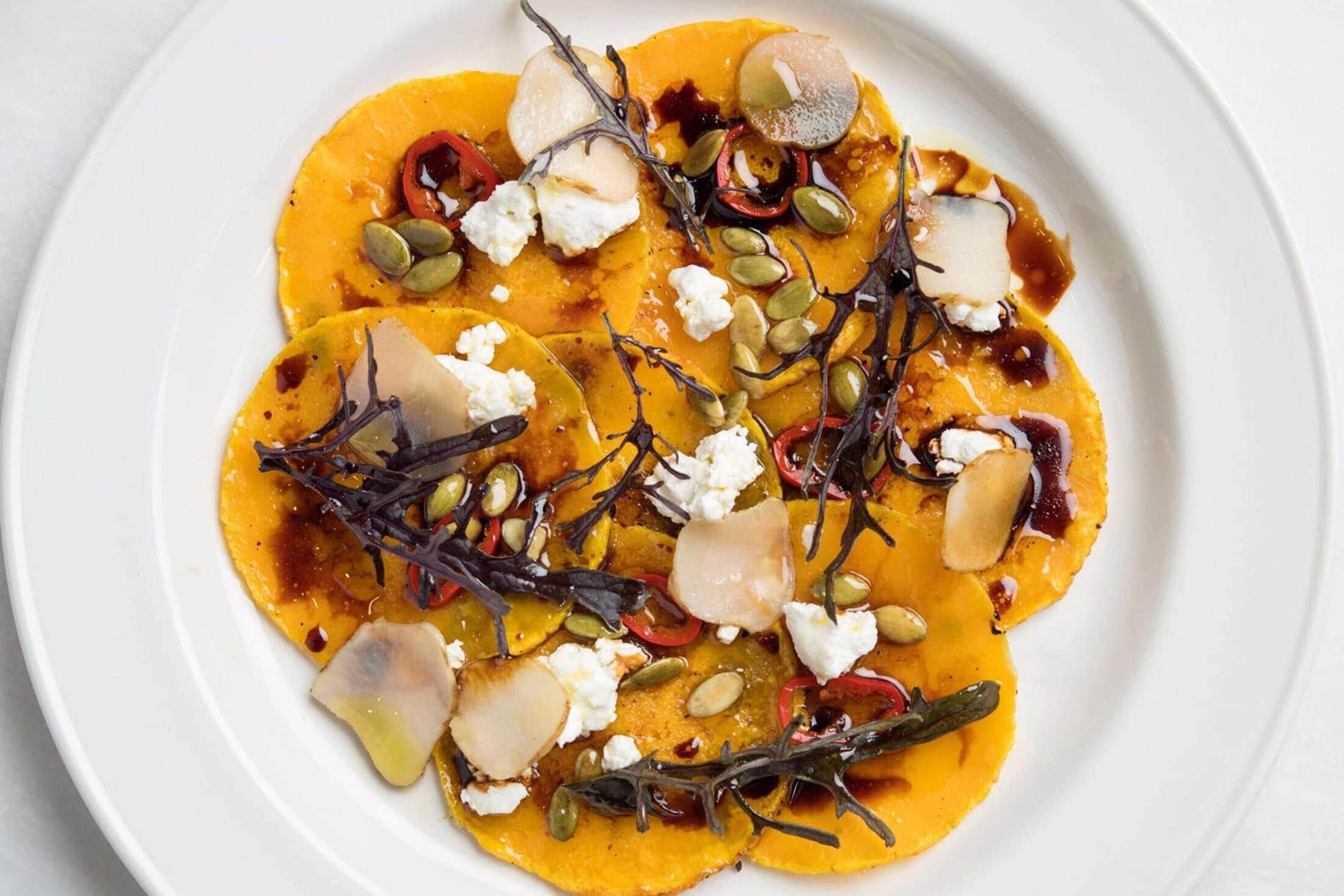 A white plate with a dish of sliced vegetables, possibly squash, topped with seeds and a drizzle of sauce.