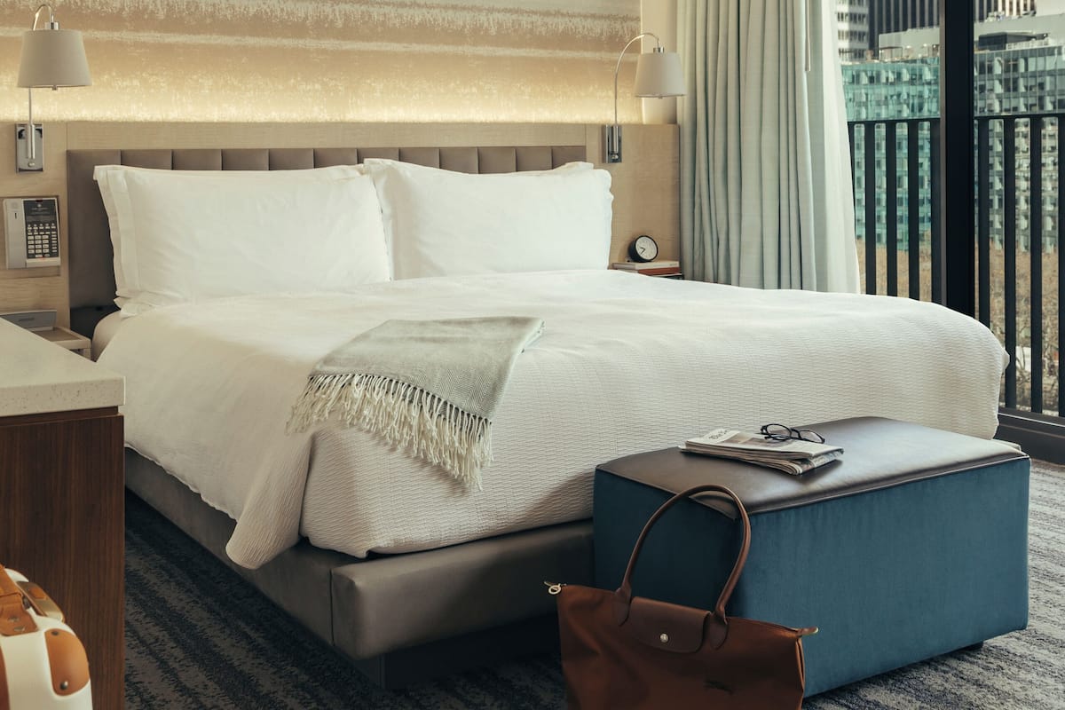 Habitación de hotel con una cama grande, una mesita de noche con lámpara, un otoman azul y vistas de la ciudad a través de la ventana.