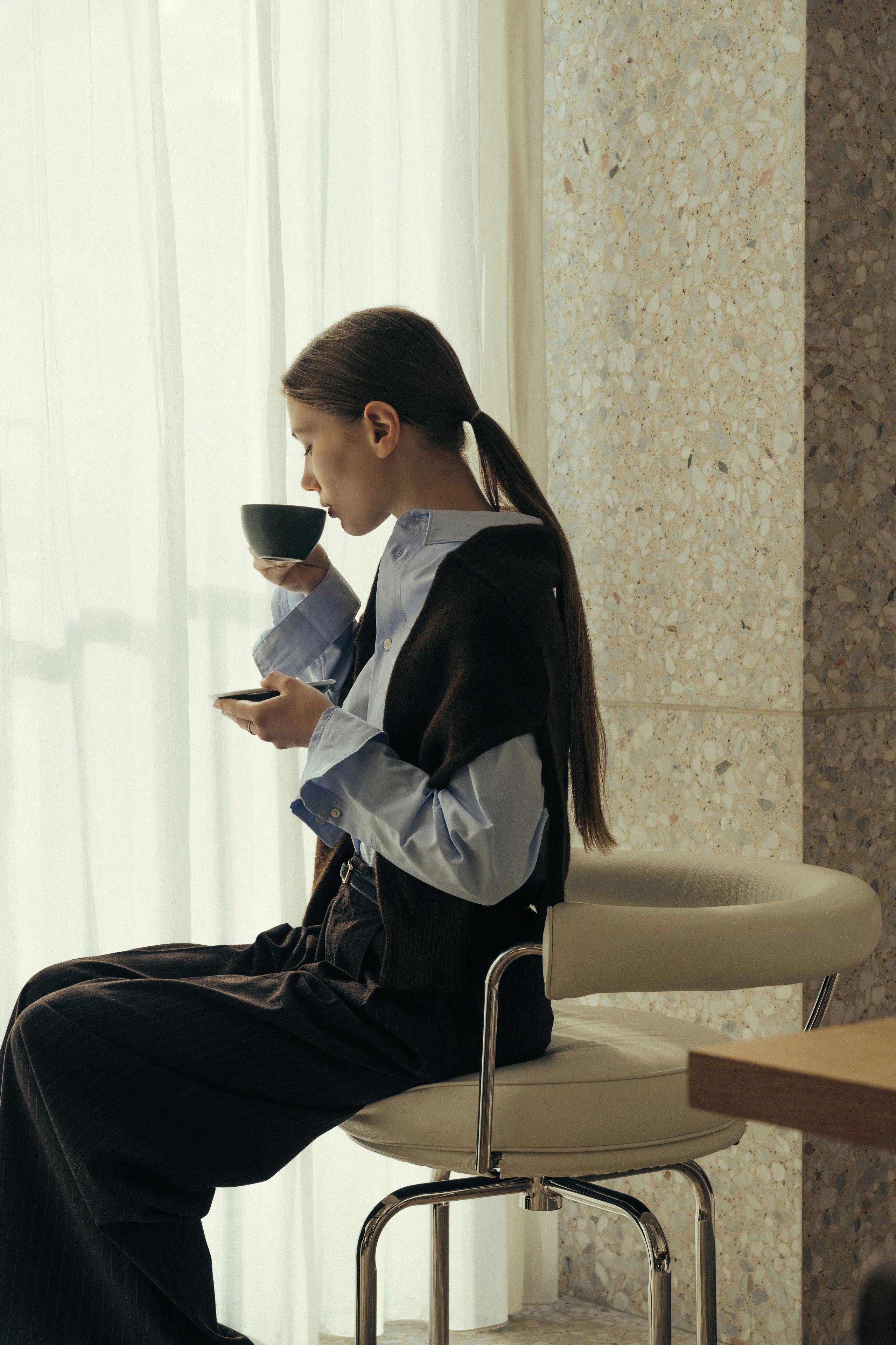 A person seated on a chair in a room with a window and a wall with a speckled pattern.