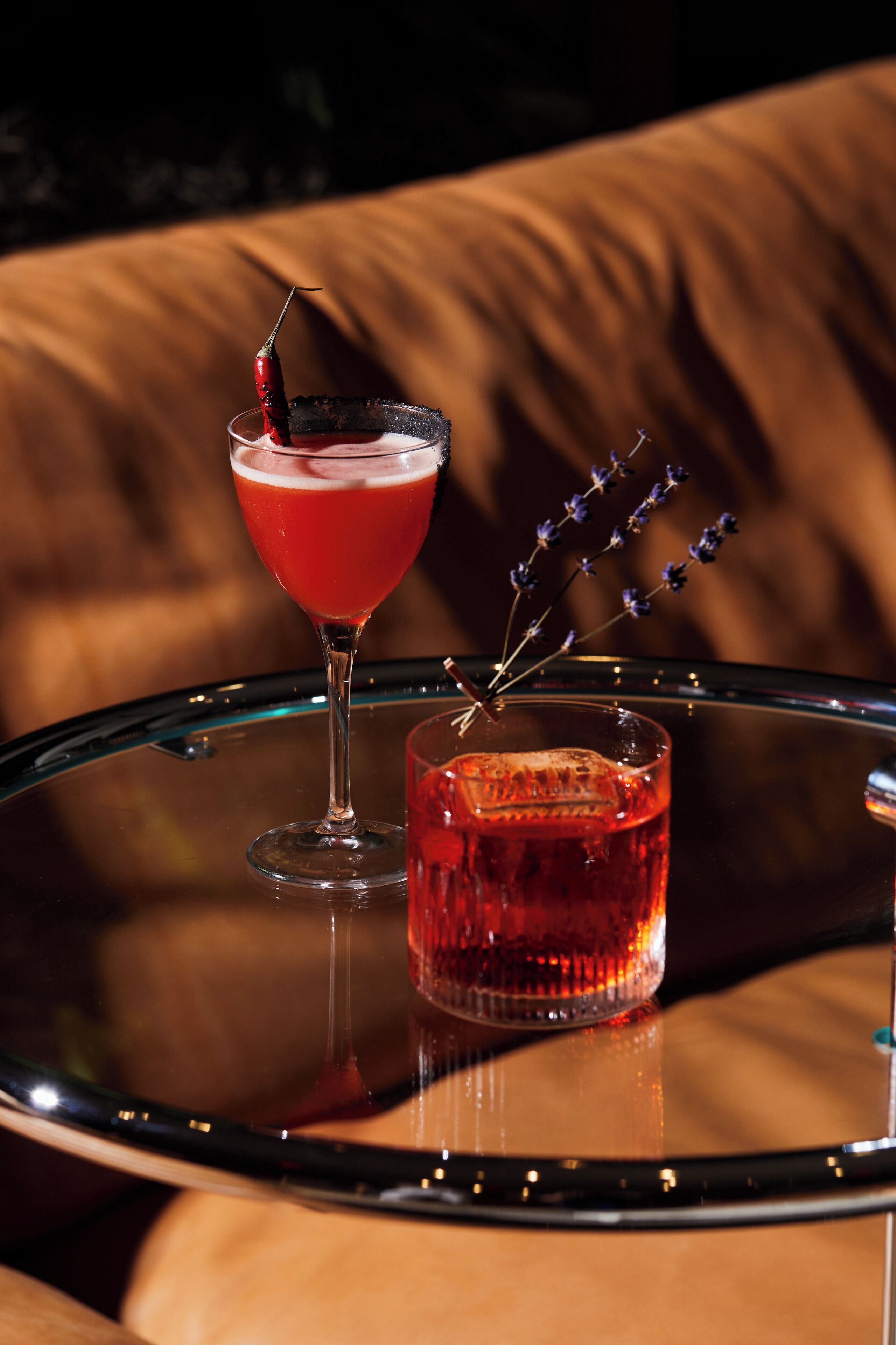 A cocktail on a glass table with a glass of red drink beside it.