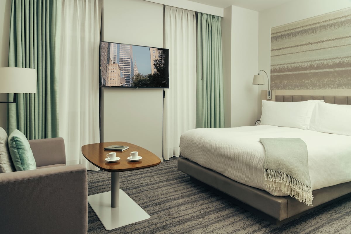 Habitación de hotel con cama, televisión montada en la pared, una mesita con una taza y un libro, y una silla.