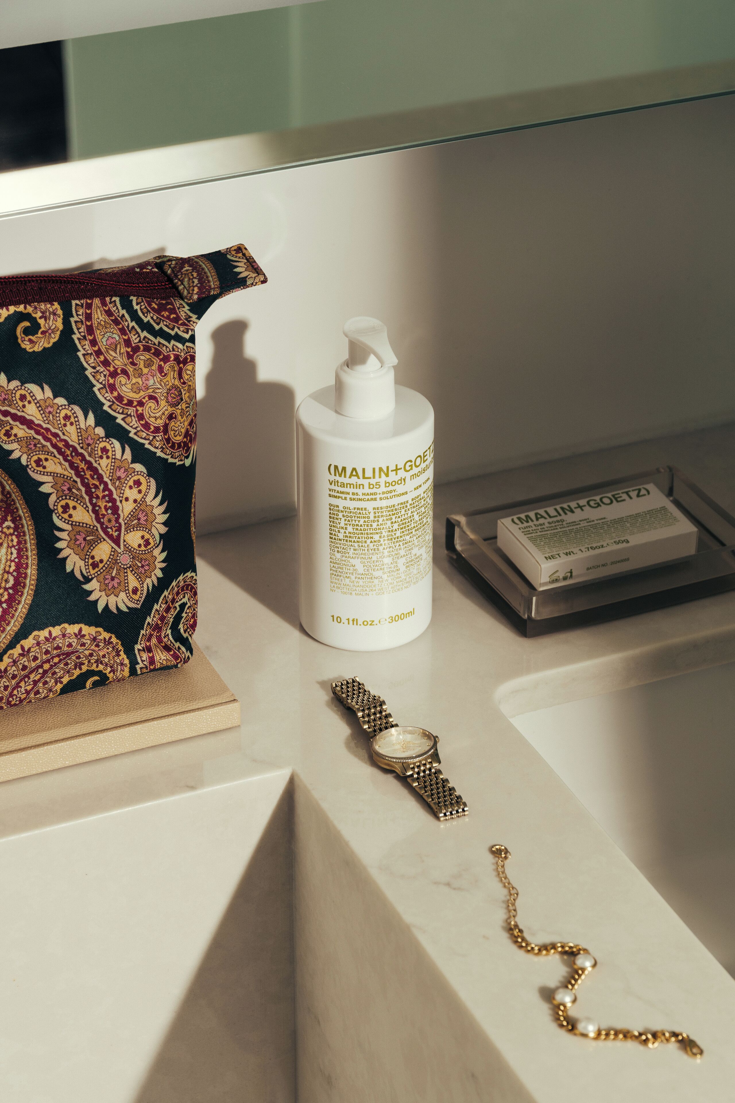 A bathroom counter with a bottle of MALIN+GOETZ body lotion, a watch, and a gold necklace.