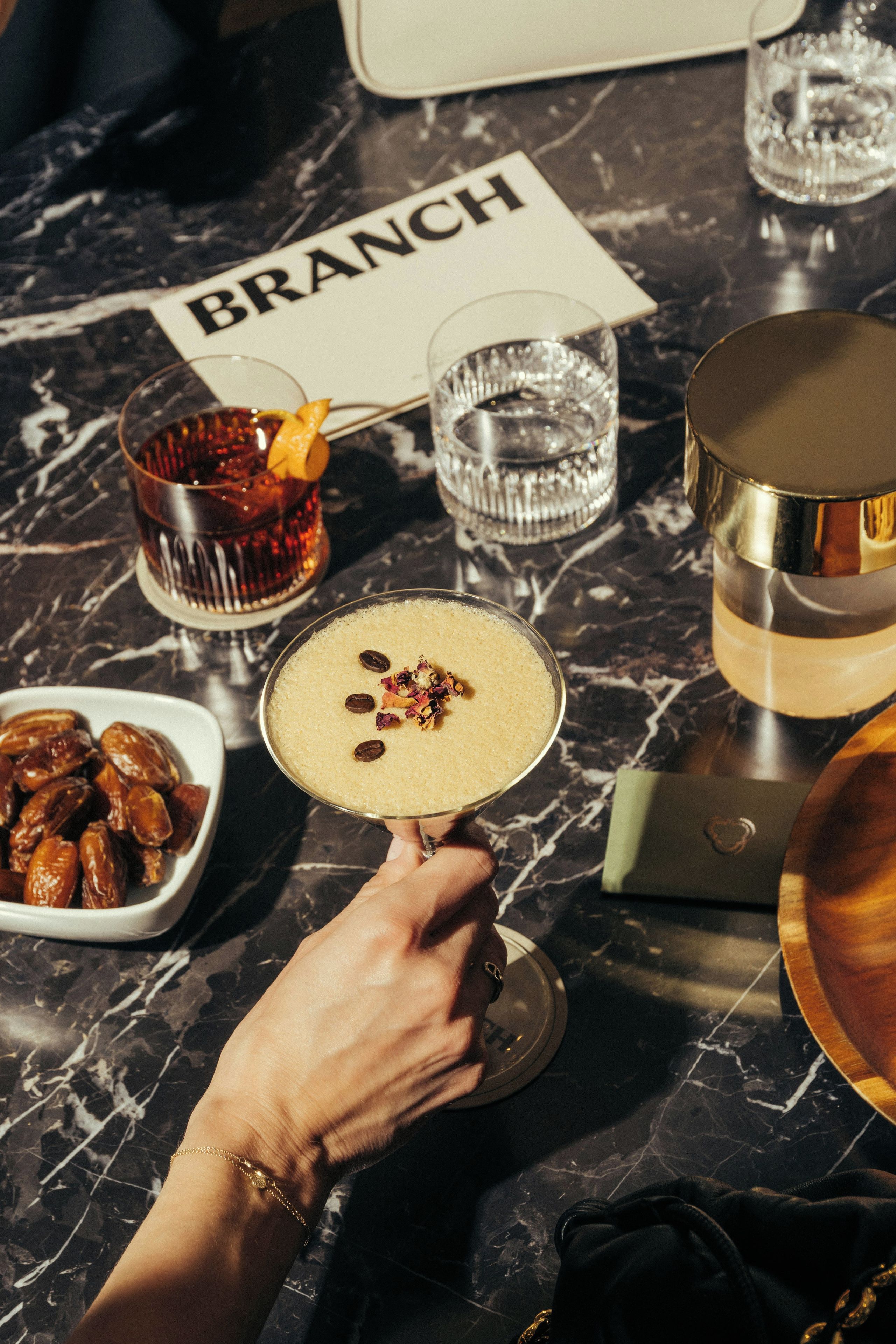 A hand holding a cocktail glass with a garnish, a bowl of dates, and a sign with the word 'BRANCH' on a marble table.