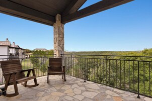 A balcony with stone flooring and wooden chairs, overlooking a green landscape.