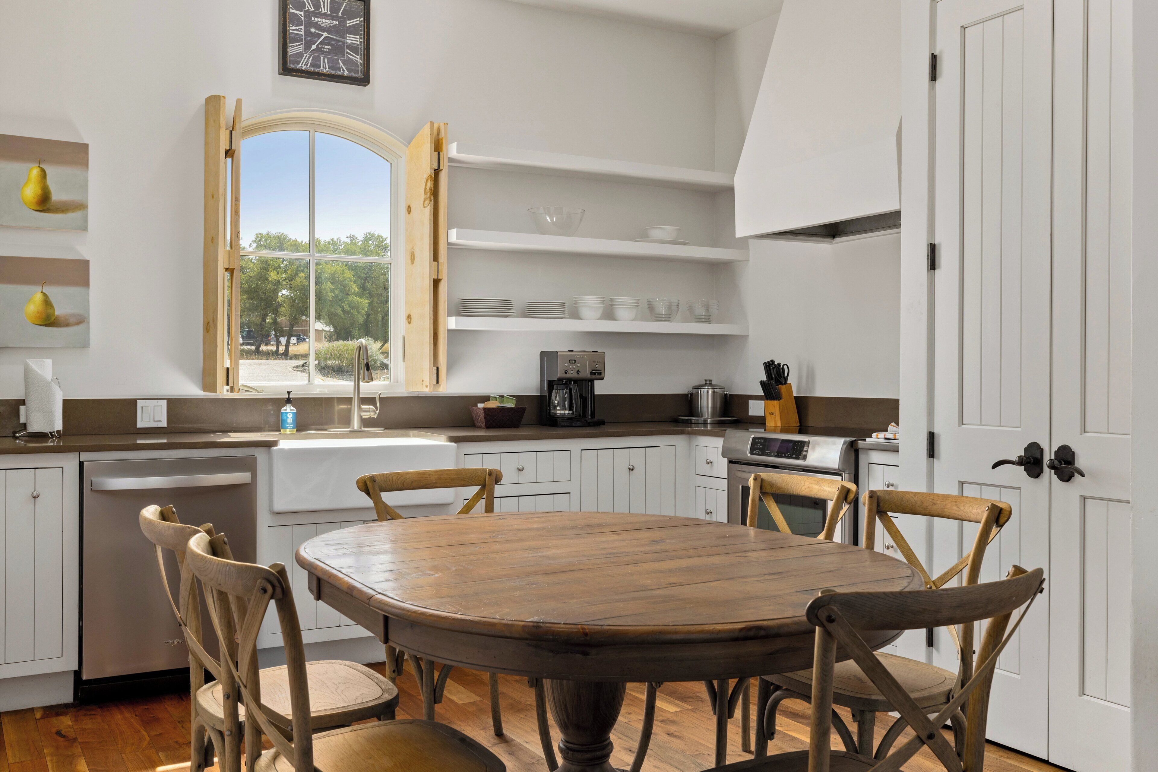 A kitchen with a round wooden table, white cabinets, and a window with an open shutter.