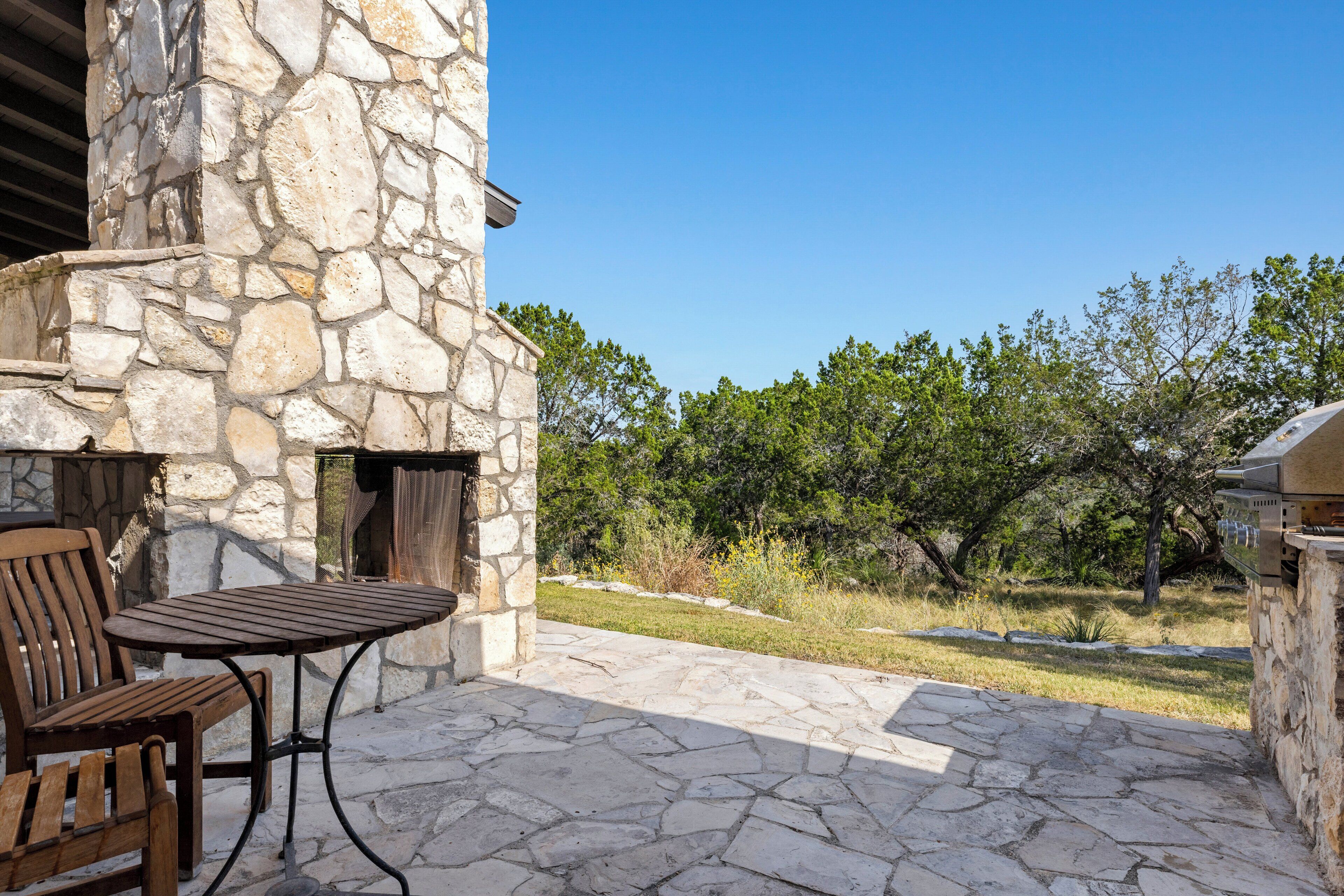 Une terrasse en pierre avec une table et des chaises, un mur en pierre et une vue sur les arbres et la pelouse.