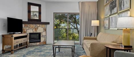 Habitación de hotel con chimenea de piedra, televisor de pantalla plana, sofá, mesa de centro y balcón con vistas a árboles.