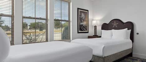 A hotel room with two beds, a large window with blinds, a framed picture on the wall, and a nightstand with a lamp.