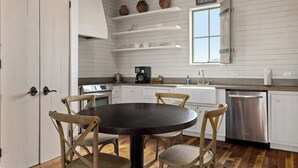 Een keuken met witte kastjes, een ronde houten tafel en vier stoelen. Boven het aanrecht zijn planken met pannen en een klok.