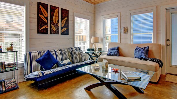 A cozy living room with a striped sofa, a glass coffee table, and a beige couch. There are two windows with blinds, a lamp on a side table, and a bookshelf filled with books. - 6 On The Beach VW6 (Port Aransas)