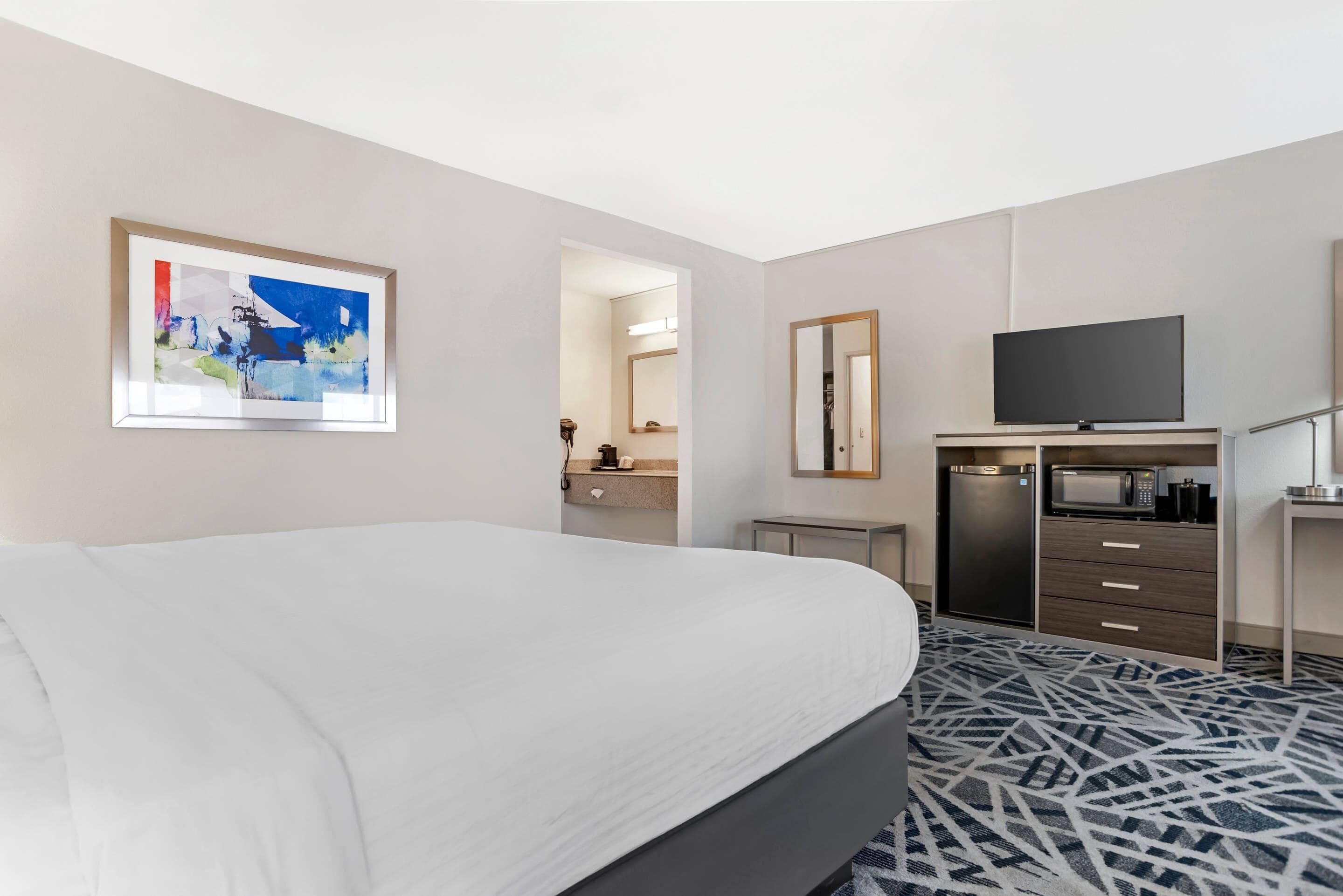 A hotel room with a bed, a television, a microwave, a refrigerator, a bathroom with a mirror, and a painting on the wall.