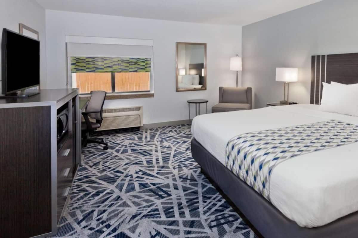 A hotel room with a bed, desk, chair, TV, and a window with blinds.