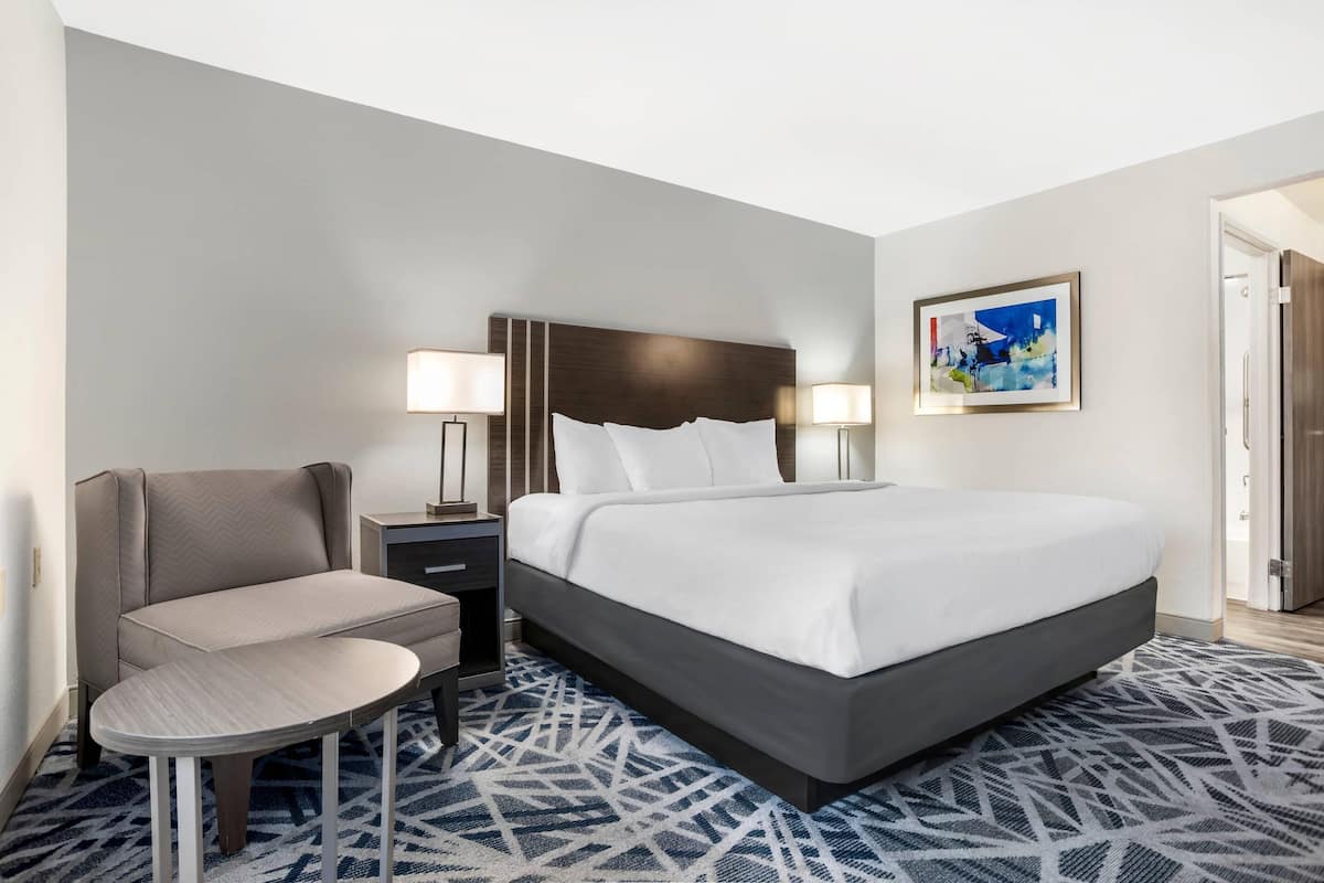 A hotel room with a bed, a chair, a desk, and a painting on the wall.