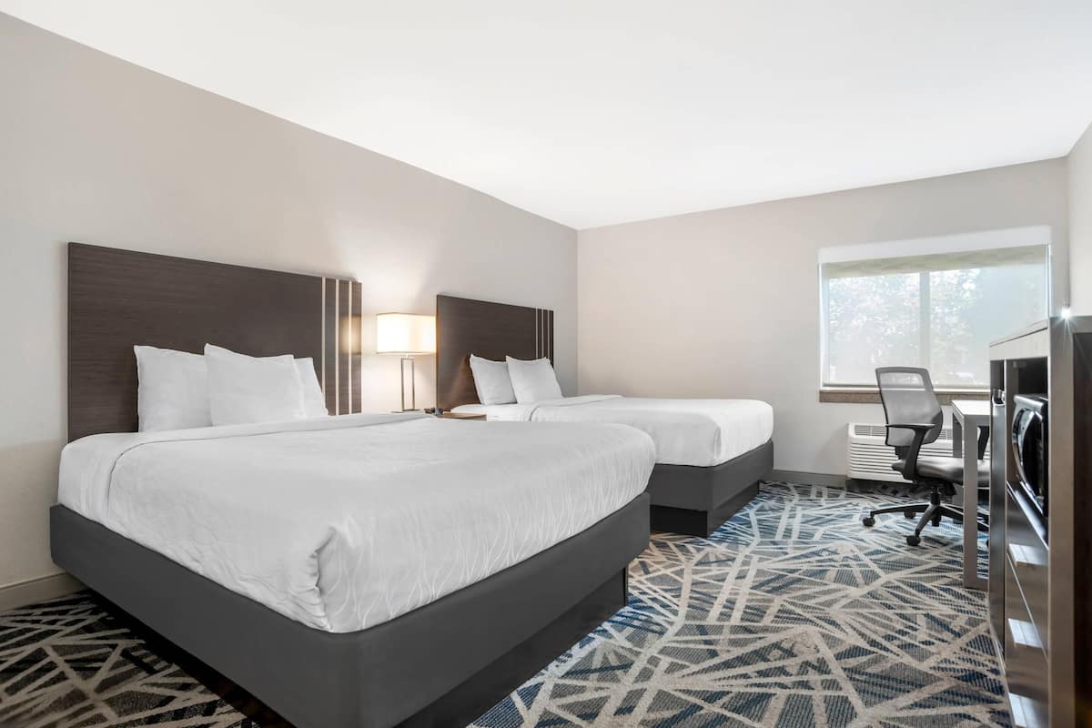 A hotel room with two beds, a desk, a chair, a lamp, and a large window.