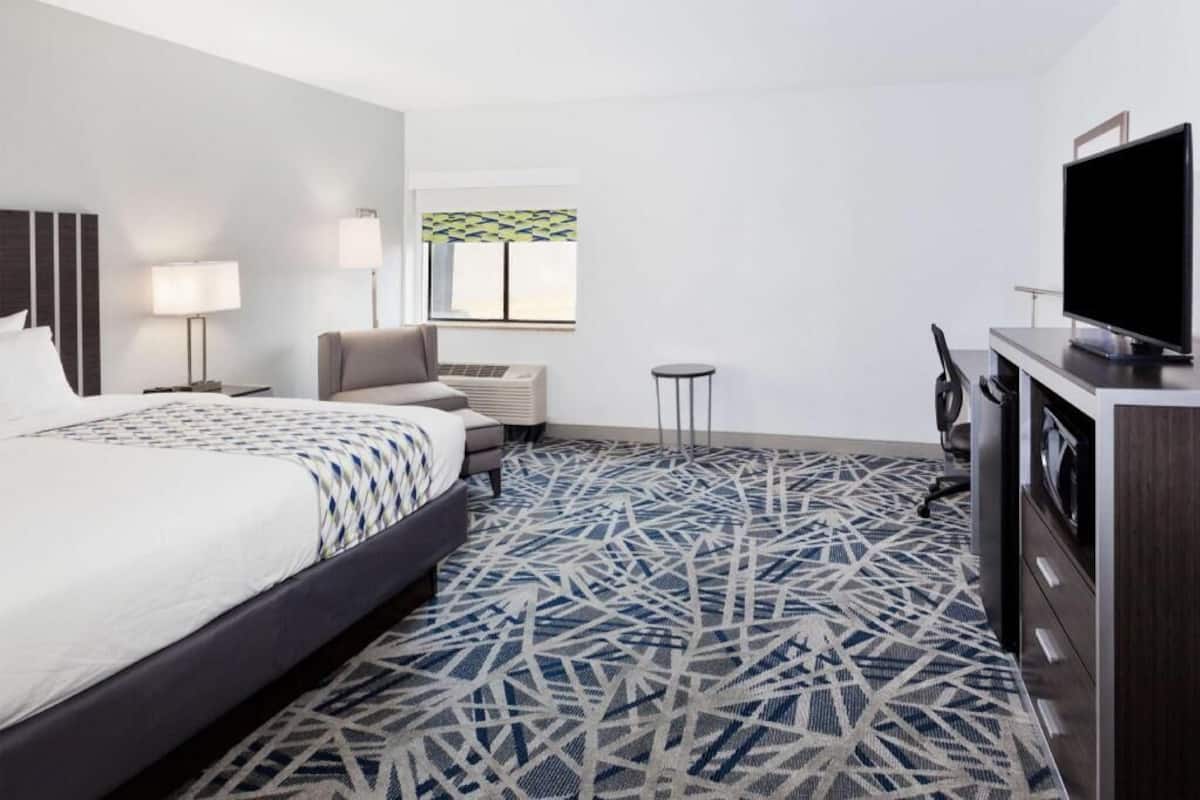 A hotel room with a bed, a desk, a television, a chair, and a window with patterned curtains.