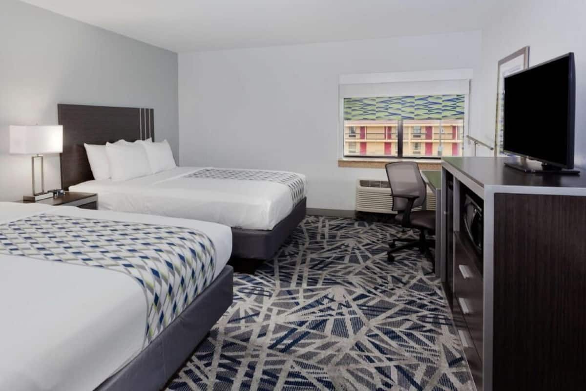 A hotel room with two beds, a desk, a TV, and a window with a view of buildings.