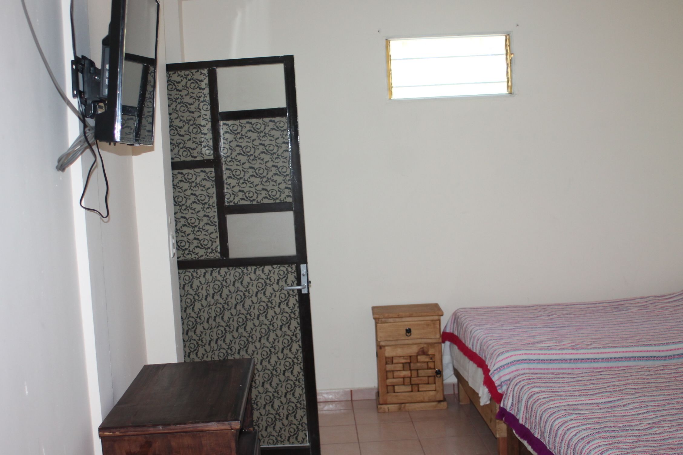 Una habitación con una cama, una mesita de noche de madera, una pequeña mesa, un televisor montado en la pared y una ventana.
